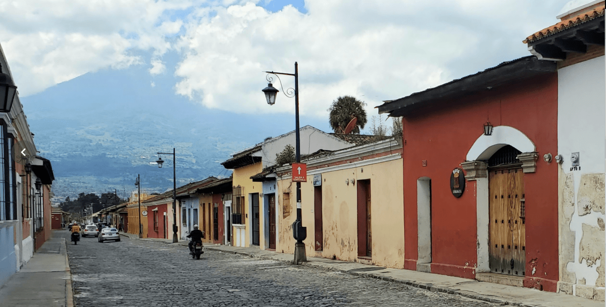 photo of a nearly empty street with one-storey buildings of different colors and colonial Spanish architecture. Hazy mountain in the background.