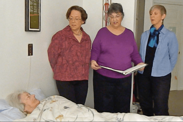 3 white women standing by an elderly white woman in bed. Their mouths are slightly open. The woman in the middle has an open binder.