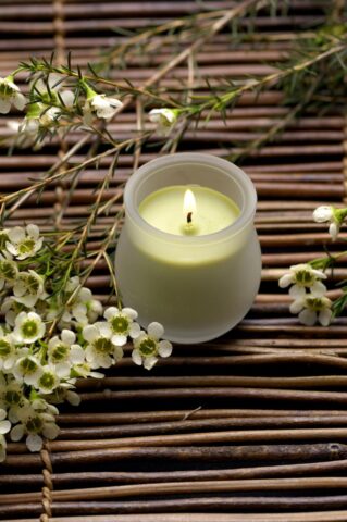 cream-colored candle in frosted glass container. Branches with small white flowers are laid next to it on a twig mat.