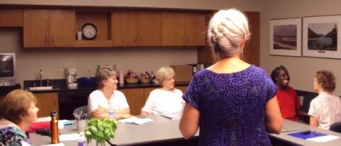 photo of women sitting around a table. In the foreground is a woman with gray hair in a French braid, facing away from the camera and toward the other women.