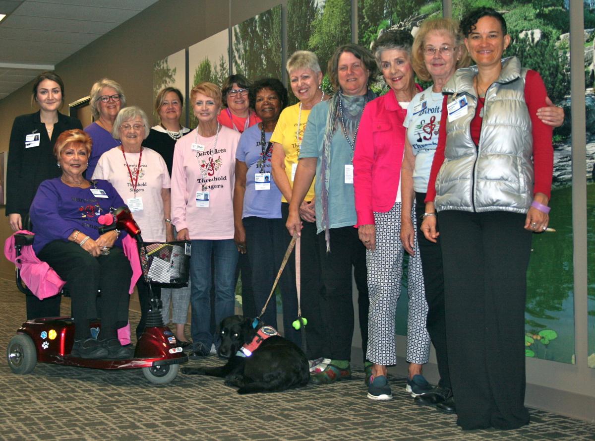 photo of 13 women, arranged by height. One woman is in a wheelchair. Some are wearing t-shirts that say "Detroit Area Threshold Singers". Several are wearing nametags on lanyards.