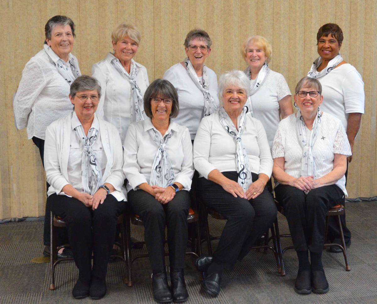 8 smiling white women and 1 brown-skinned woman. All are wearing white shirts and scarves with music symbols on them