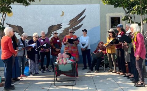 group of woman standing outdoors, singing to a person in a reclining chair