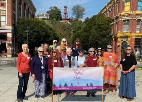 Group of women standing in a downtown location, holding a Threshold Choir banner