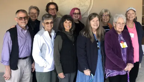 Group of smiling mostly older white men and women. Two women has a metal cane. They are dressed in vests and jackets.