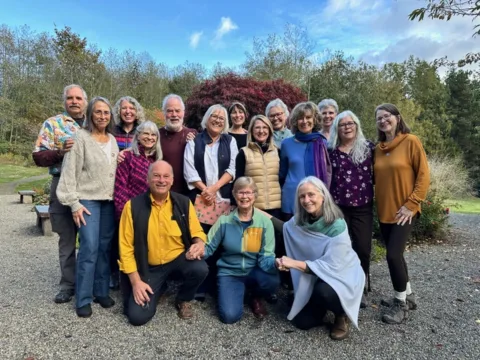 16 smiling white people outdoors on gravel, trees in the background