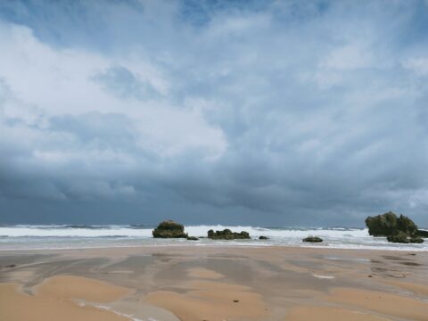 Wet sand in foreground, water with some rocks in the background. Sky is blue and cloudy.