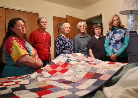 6 white women standing around a bed with a red, white, and blue quilt